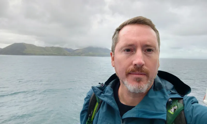 a white man with dark eyes and a white trimmed beard takes a selfie with the ocean and some hills behind him.