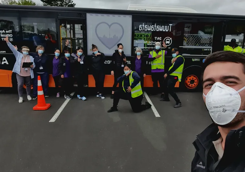 A man wearing a face masks takes a selfie in front of a vaccination bus and a group of people wearing masks and hi-vis