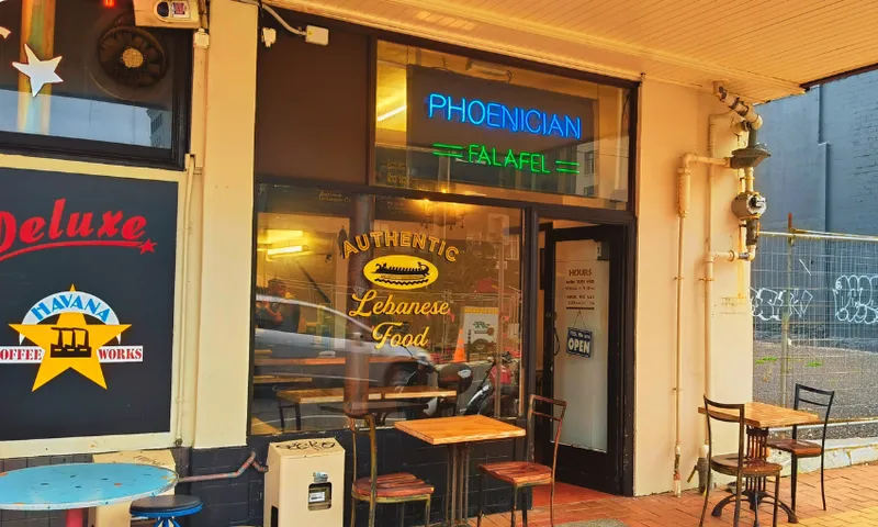 Street view of a small restaurant with signs reading "PHOENICIAN FALAFEL" in neon and "AUTHENTIC Lebanese Food" on the window. Outdoor tables and chairs line the brick sidewalk. A "Havana Coffee Works" sign is visible on the left.