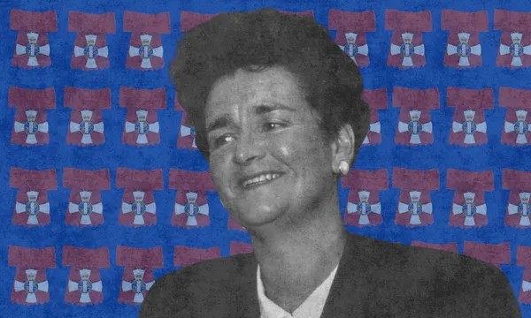 A black-and-white photo of a woman in a suit, smiling, set against a blue background patterned with red and blue flags featuring a central emblem.
