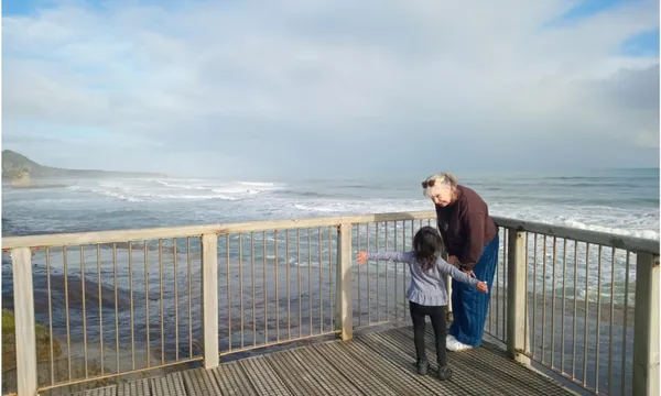A photo of an older woman standing on a pier with a young girl. They are talking to each other. There is a vast sky with clouds.