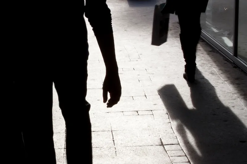 Two people walk on a sunlit sidewalk, casting long shadows. One person is closer to the camera with only part of their silhouette visible, while the other walks away holding a bag. The scene is contrasted with strong lighting.