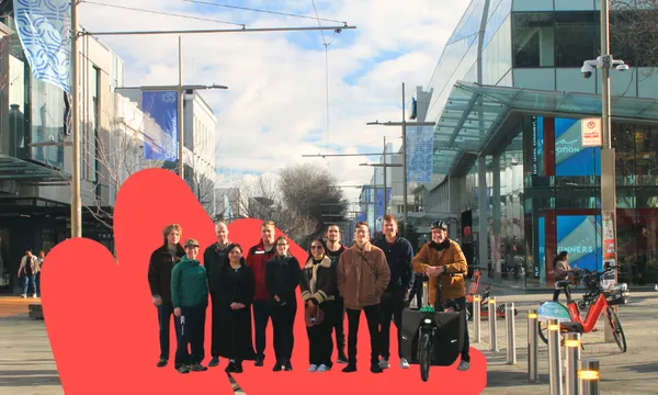 a group of people standing on a christchurch street with a red squiggle behind them. several have bikes; all are smiling but look a bit cold.