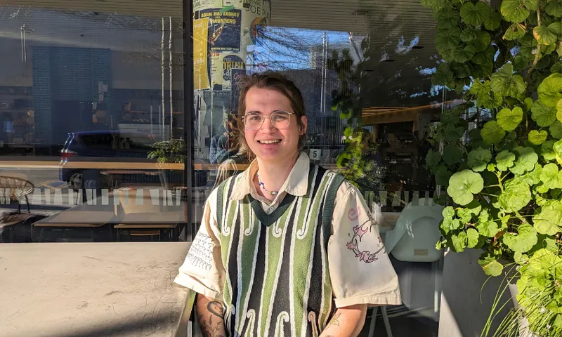 a person sitting in the sunshine at a table in a green and black vest with a white button up shirt underneath smiling