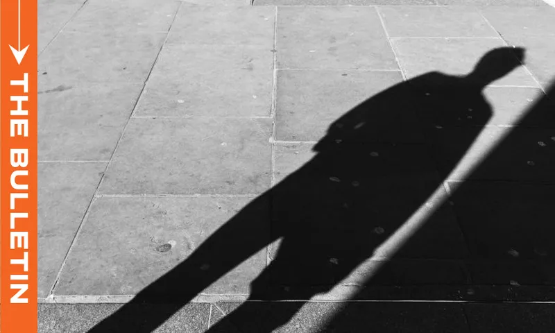 A long shadow of a person is cast on a sunlit pavement. On the left, an orange vertical banner reads "THE BULLETIN" in white text, with a white arrow above it.