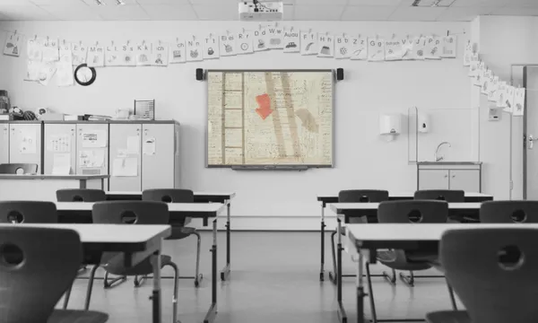 A black-and-white classroom with empty desks faces a smartboard displaying a sepia-toned handwritten document, the Treaty of Waitangi. Children’s drawings hang along the wall above white cabinets.