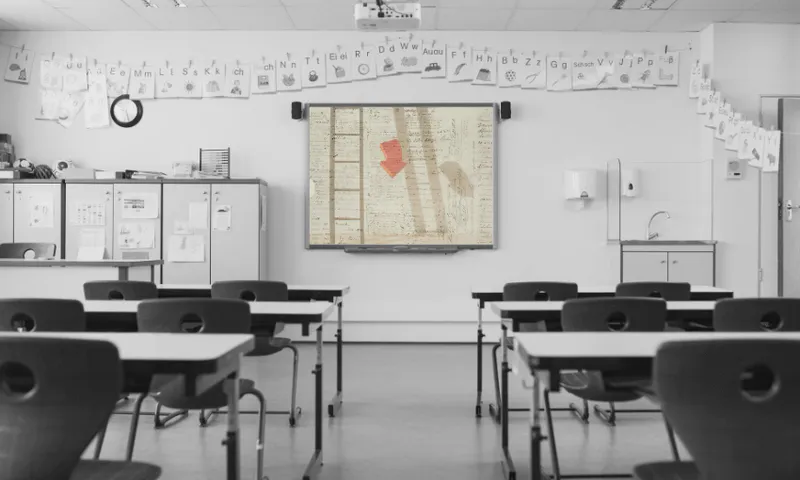 A black-and-white classroom with empty desks faces a smartboard displaying a sepia-toned handwritten document, the Treaty of Waitangi. Children’s drawings hang along the wall above white cabinets.