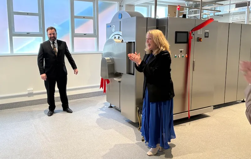 A man in a suit and a woman in a blue dress clap hands next to a large silver machine