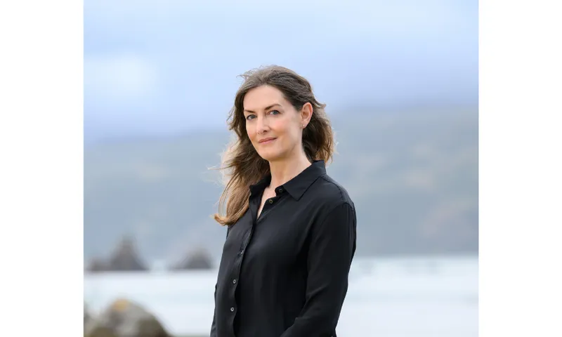 Photograph of a woman with long brown hair standing outside near a beach. She is looking at the camera.