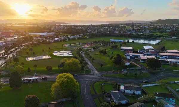 Aerial view of a suburban neighborhood at sunset, featuring green parks, sports fields, scattered houses, and buildings with trees and roads throughout the landscape. The sun is low on the horizon, casting a warm light.