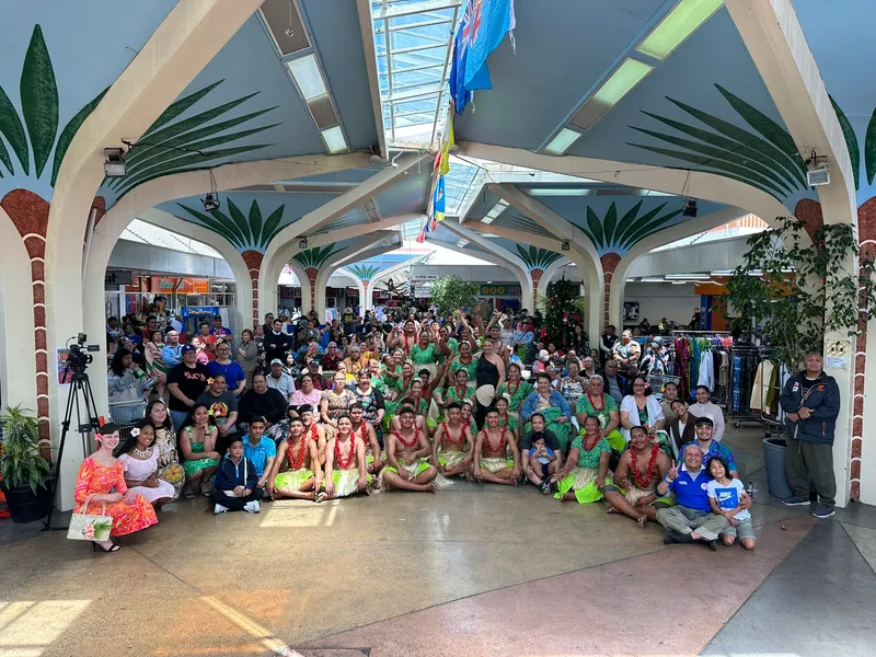 A large group of people, including dancers in traditional Hawaiian attire, pose for a photo in a decorated indoor space with palm tree pillars. The scene is lively and colorful, with various expressions of joy and camaraderie.