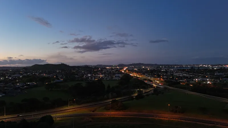 A cityscape at dusk with a winding road lit by car headlights, green fields in the foreground, and scattered houses. The sky is transitioning from blue to night with clouds and distant hills on the horizon.