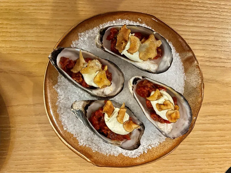 Four oyster shells filled with a raw beef mixture, garnished with thin, crispy chips, are arranged on a bed of coarse salt on a brown ceramic plate atop a wooden table.