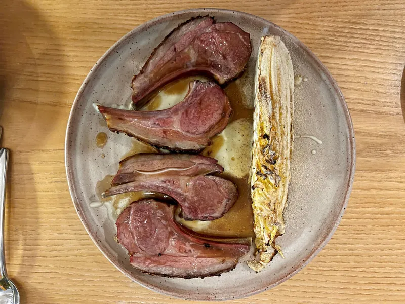 A plate with four cooked lamb chops arranged in a semicircle and a piece of grilled endive, all served on a light brown ceramic plate on a wooden table.