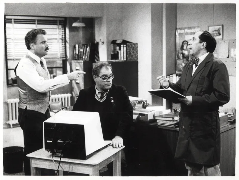 Three men in an office: one standing and pointing, one sitting at a desk with a computer, and another standing with a clipboard, engaged in conversation. The room has shelves, books, and office equipment.