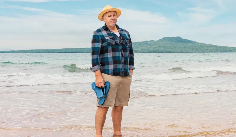 An older man stands barefoot on a beach, holding blue sandals. He wears a straw hat, plaid shirt, and khaki shorts. Gentle waves and a green, hilly island are visible in the background under a partly cloudy sky.