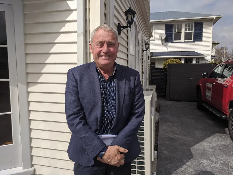 an image of an older white man in a blue suit smiling while holding a folder in front of a weatherboard house