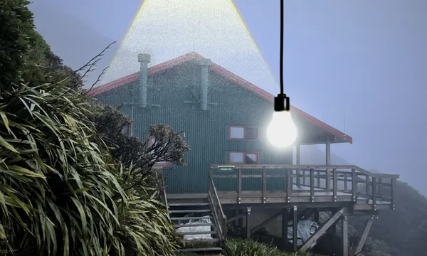 A green backcountry hut set against the side of a mountain on a foggy night. An electric lightbulb hangs down from the top of the frame