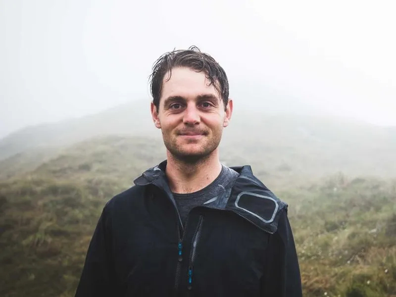A man with short brown hair and a slight smile stands outdoors on a foggy day, wearing a black jacket. Grassy hills are faintly visible in the misty background.