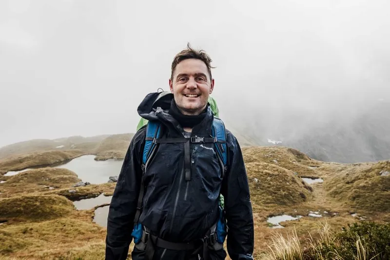A person wearing a black rain jacket and backpack smiles while standing outdoors in a foggy, grassy landscape with small ponds and rolling hills in the background.