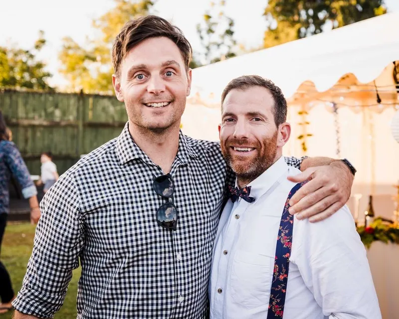 Two men smiling outdoors at an event. One has his arm around the other; he wears a checked shirt with sunglasses hanging from the collar. The other wears a white shirt, floral suspenders, and a bow tie. Tents and trees are in the background.