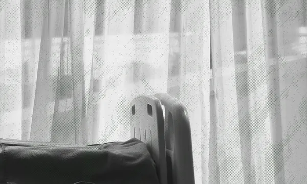A black and white photo of a hospital bed partially visible in front of sheer curtains, with soft daylight coming through the window.