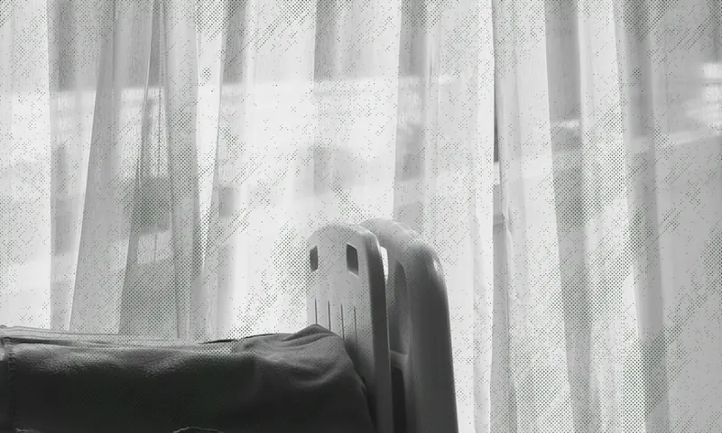 A black and white photo of a hospital bed partially visible in front of sheer curtains, with soft daylight coming through the window.