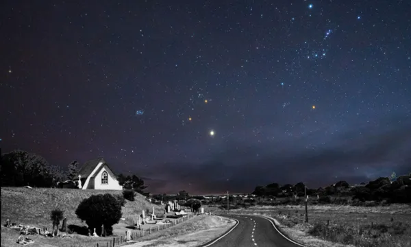 A rural road curves past a small house and gravestones under a clear, star-filled night sky with visible constellations. The landscape is dimly lit, creating a peaceful, serene atmosphere.