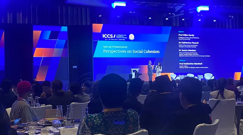A faraway photo of a conference stage in a dimly lit function room, with a large blue screen at the back and two speakers on the stage