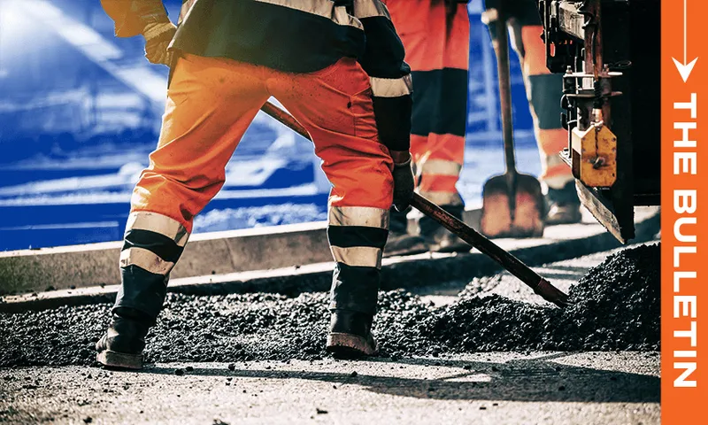 Two road workers in high-visibility orange pants use shovels to spread asphalt on a road construction site. "The Bulletin" is written vertically on the right side of the image.
