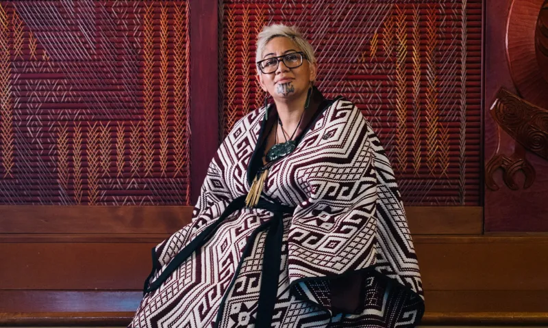 A person with glasses and traditional facial markings sits wrapped in a patterned cloak, in front of a wall with intricate wood carvings and patterns.