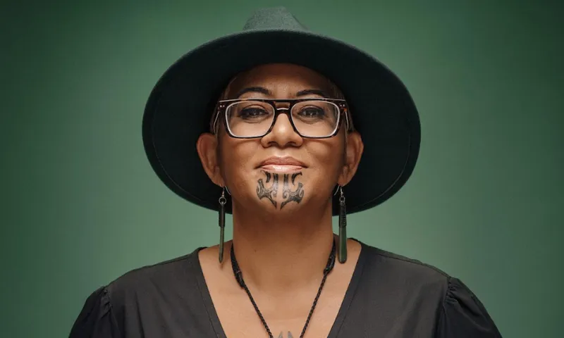 A person wearing a wide-brimmed dark hat, glasses, and earrings, with a traditional Māori chin tattoo and a confident expression, poses against a solid green background.