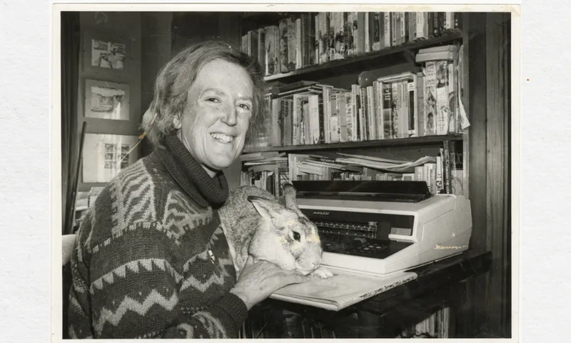 A black and white photo of the writer Margaret Mahy. She is sitting in a room with a lot of bookshelves. She's sitting at a desk with a typewriter in front of her, and a large rabbit on her knees. She is smiling.