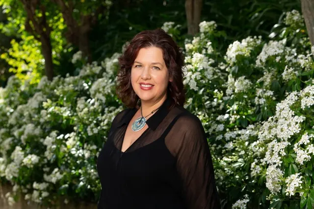 a white woman in a black top with a floral bush behind her