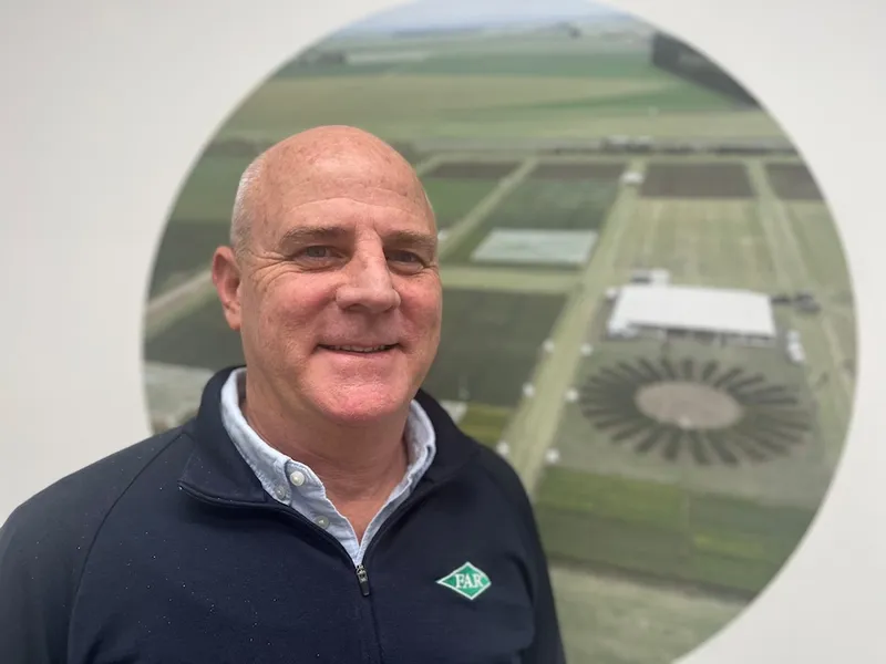 a bald man with pale skin in front of a photo of green fields