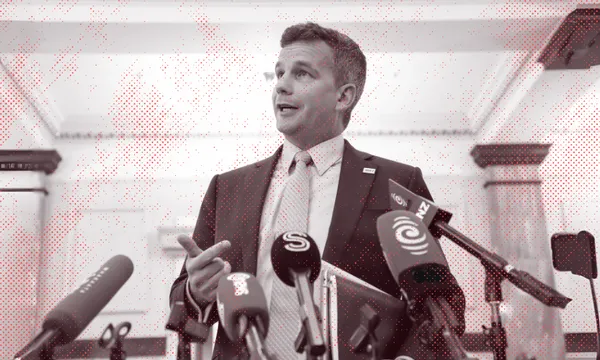 A man in a suit speaks at a press conference, standing behind several microphones from various news outlets in a formal indoor setting.