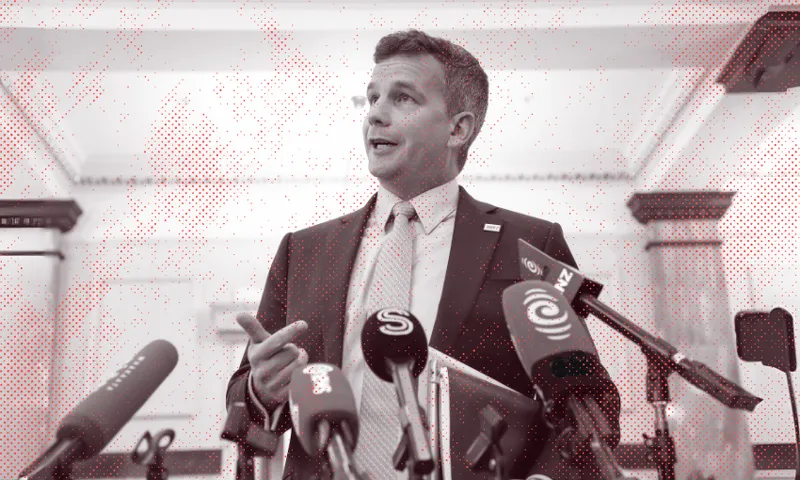 A man in a suit speaks at a press conference, standing behind several microphones from various news outlets in a formal indoor setting.