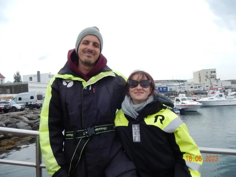 a young man and woman smile with ice winter gear on. there is water in the background and they are wearing beanies and sunglasses