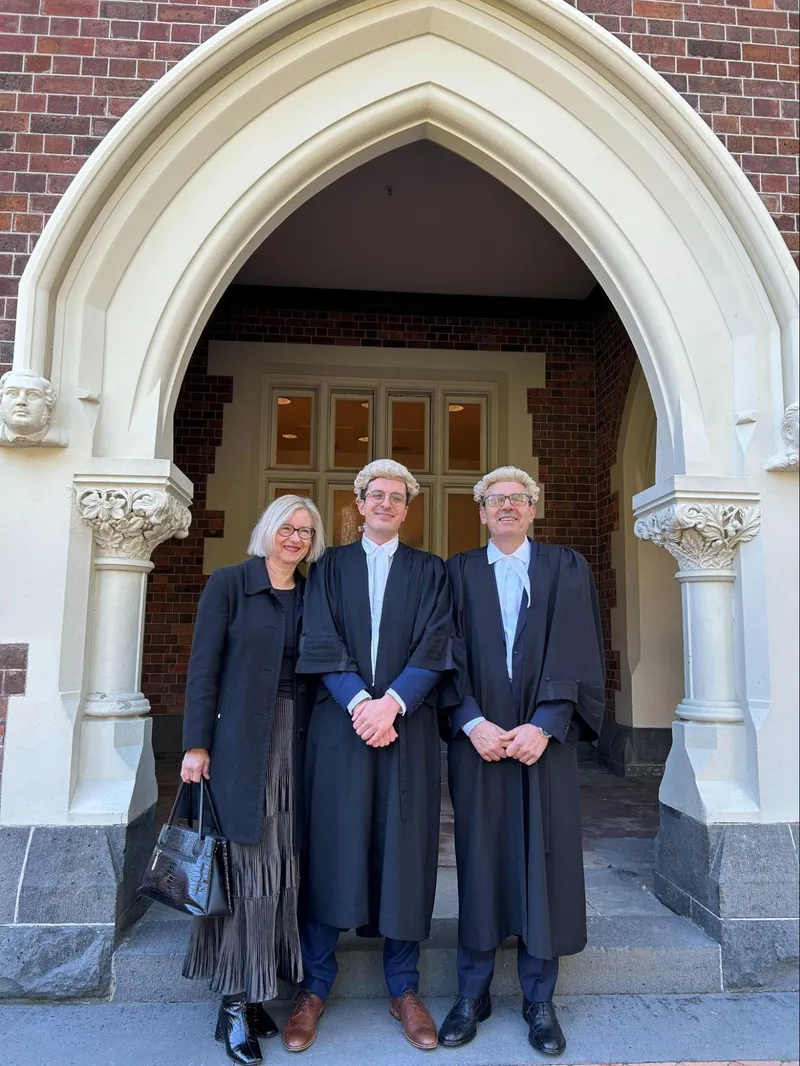 a young man stands with his parents. he and his father wear black law robes and white wigs, his mother wears a black blazer and skirt