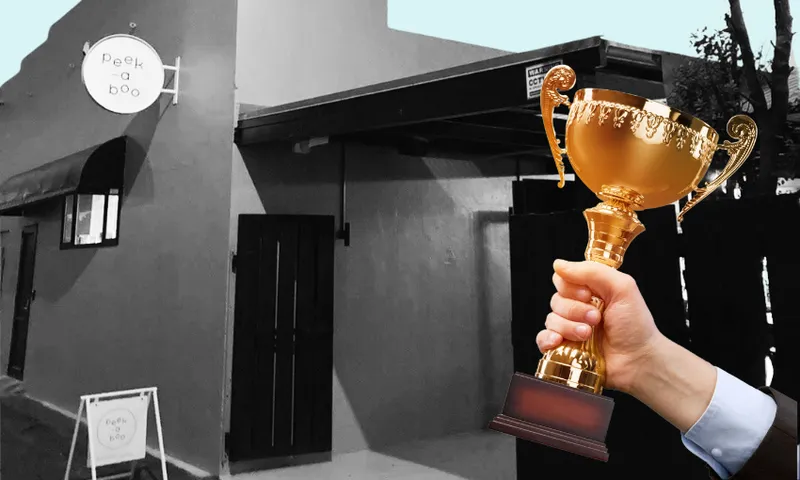 A hand holding a gold trophy in front of a building with a round "Peek-a-boo" sign; the image is mostly black and white except for the trophy, which is in color.