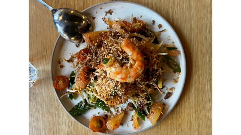 A plate of salad topped with a large shrimp, shredded vegetables, cherry tomatoes, and crispy toppings is served on a wooden table next to a metal spoon.