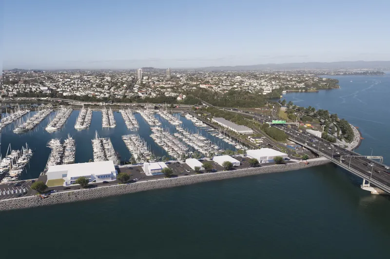 Aerial view of a marina filled with boats next to a coastal cityscape, with buildings, roads, and a bridge extending over the water, bordered by trees and surrounded by calm blue water.