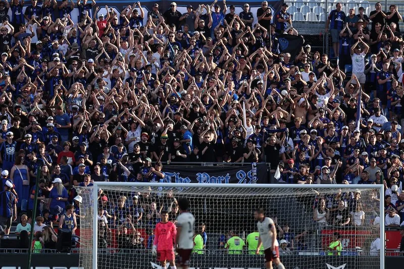 A large crowd of enthusiastic soccer fans in blue shirts cheer and raise their arms in unison in the stadium stands, while players and a goalkeeper are visible near the goal on the field.