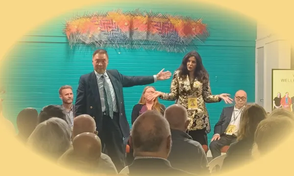 Two presenters stand in front of a seated audience, gesturing expressively. Three other panelists are seated behind them. The background features a teal wall and colorful artwork. The scene has a soft yellow vignette effect.