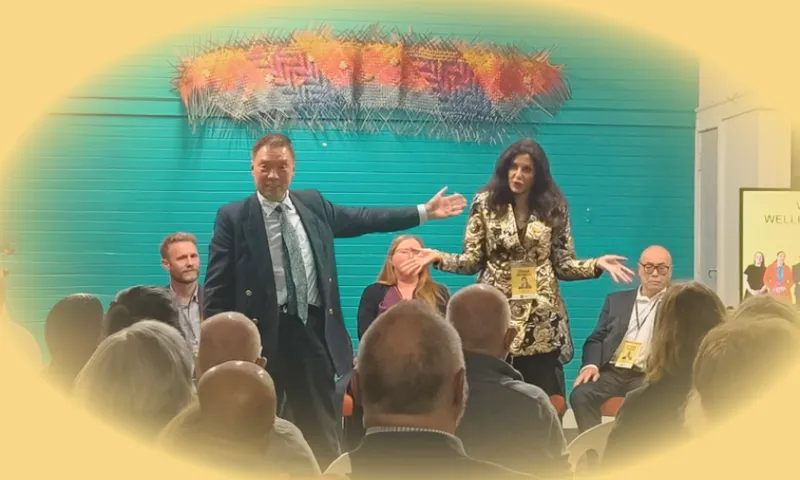 Two presenters stand in front of a seated audience, gesturing expressively. Three other panelists are seated behind them. The background features a teal wall and colorful artwork. The scene has a soft yellow vignette effect.