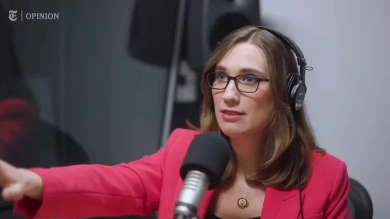 A woman wearing glasses and headphones speaks into a microphone in a recording studio. She is wearing a pink blazer and gesturing with her hand.
