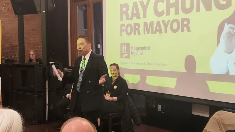 A man in a suit speaks in front of an audience at an indoor event. Behind him, a large yellow screen displays the text “Ray Chung for Mayor” and “independent together.” Several people are seated nearby.