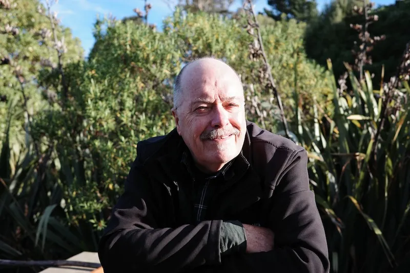 a man smiling in the sun, with white hair and a brown jacket, and trees in the background