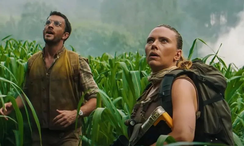 A still from the film Jurassic Park: Rebirth showing the actors Jonathan Bailey and Scarlett Johansson standing in shoulder-high grass and looking up at something that is shocking them.