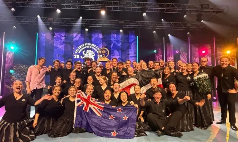 A group photo of the 2025 youth choir. About 40 young people wearing black and silver uniforms and smiling while holding a trophy and a New Zealand flag